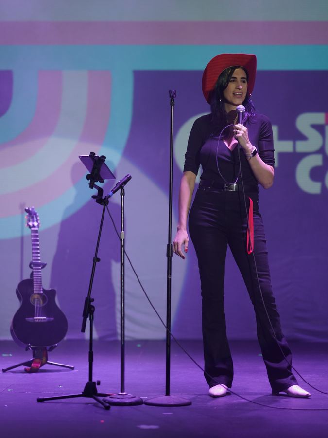 Ophelia con sombrero vaquero y guitarra en el escenario de Q+ Comedia Diversa, luz morada. Vertical.