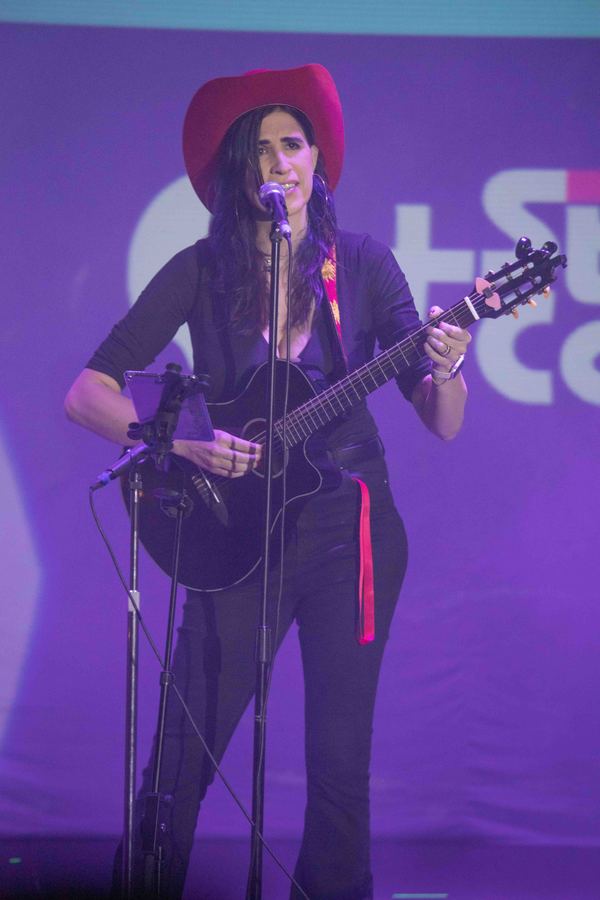 Ophelia cantando con guitarra y sombrero vaquero, escenario morado. Vertical.