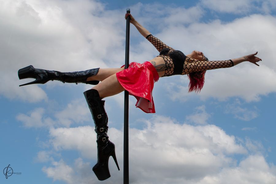 Ophelia extendida en horizontal sobre un tubo de pole al aire libre, cielo con nubes detrás, botas negras y falda roja. Horizontal.