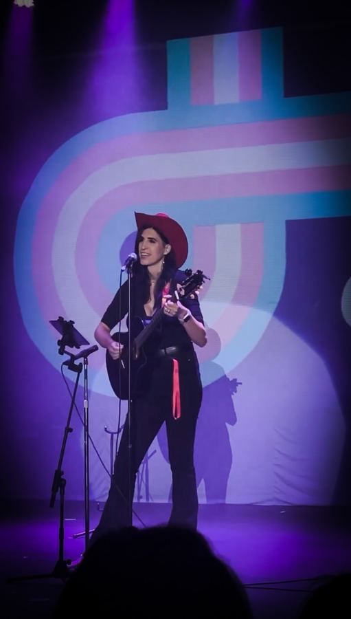 Ophelia con sombrero y guitarra en un escenario, luz morada y rosa. Vertical.