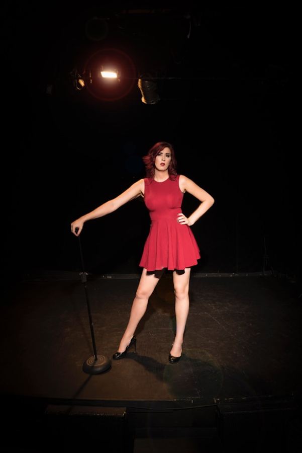 Ophelia con vestido rojo en un escenario de stand up, mano sobre el micrófono.