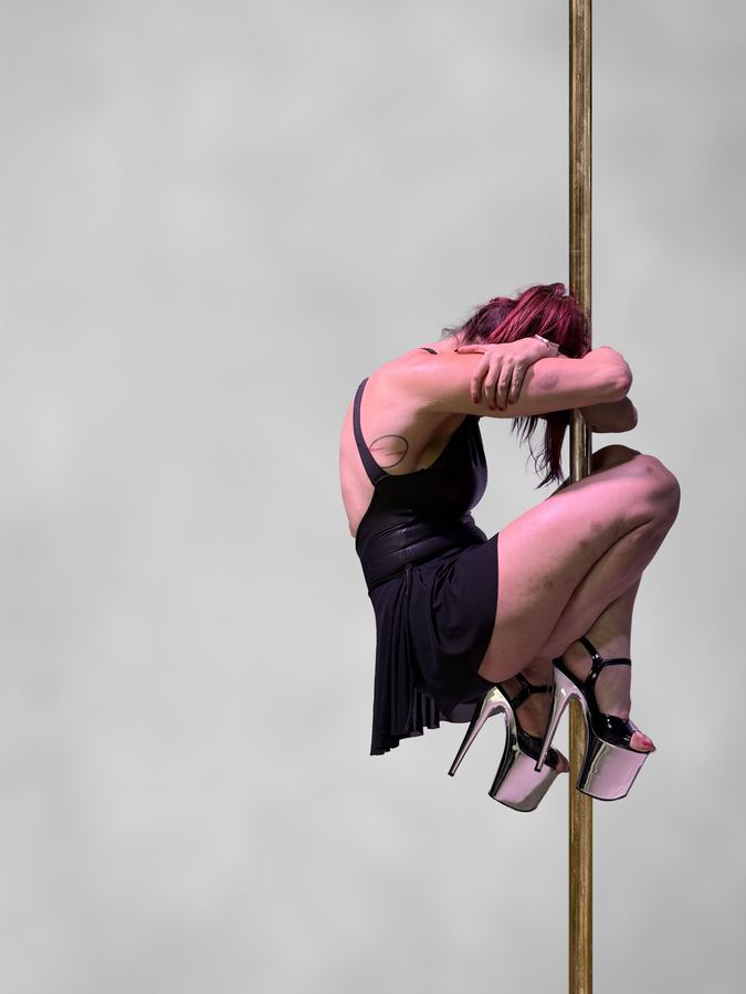 Ophelia abrazada al tubo de pole con vestido rosa, fondo claro al aire libre. Vertical.