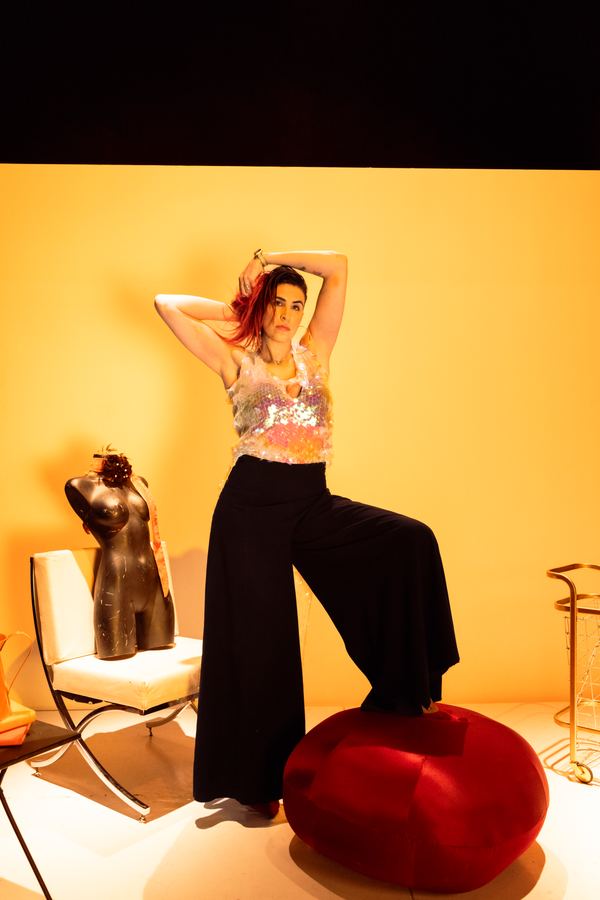 Ophelia de pie con pantalón negro ancho, brazo sobre la cabeza, fondo amarillo.