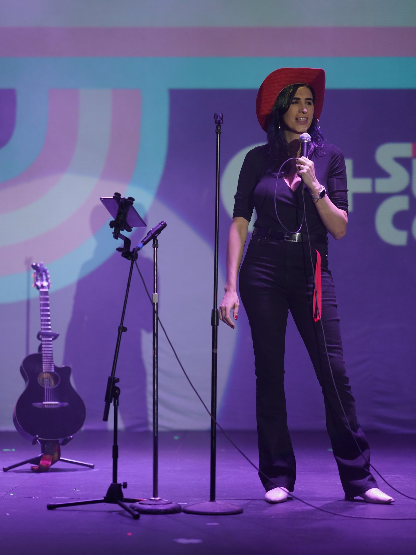 Ophelia con sombrero vaquero y guitarra en el escenario de Q+ Comedia Diversa, luz morada. Vertical.
