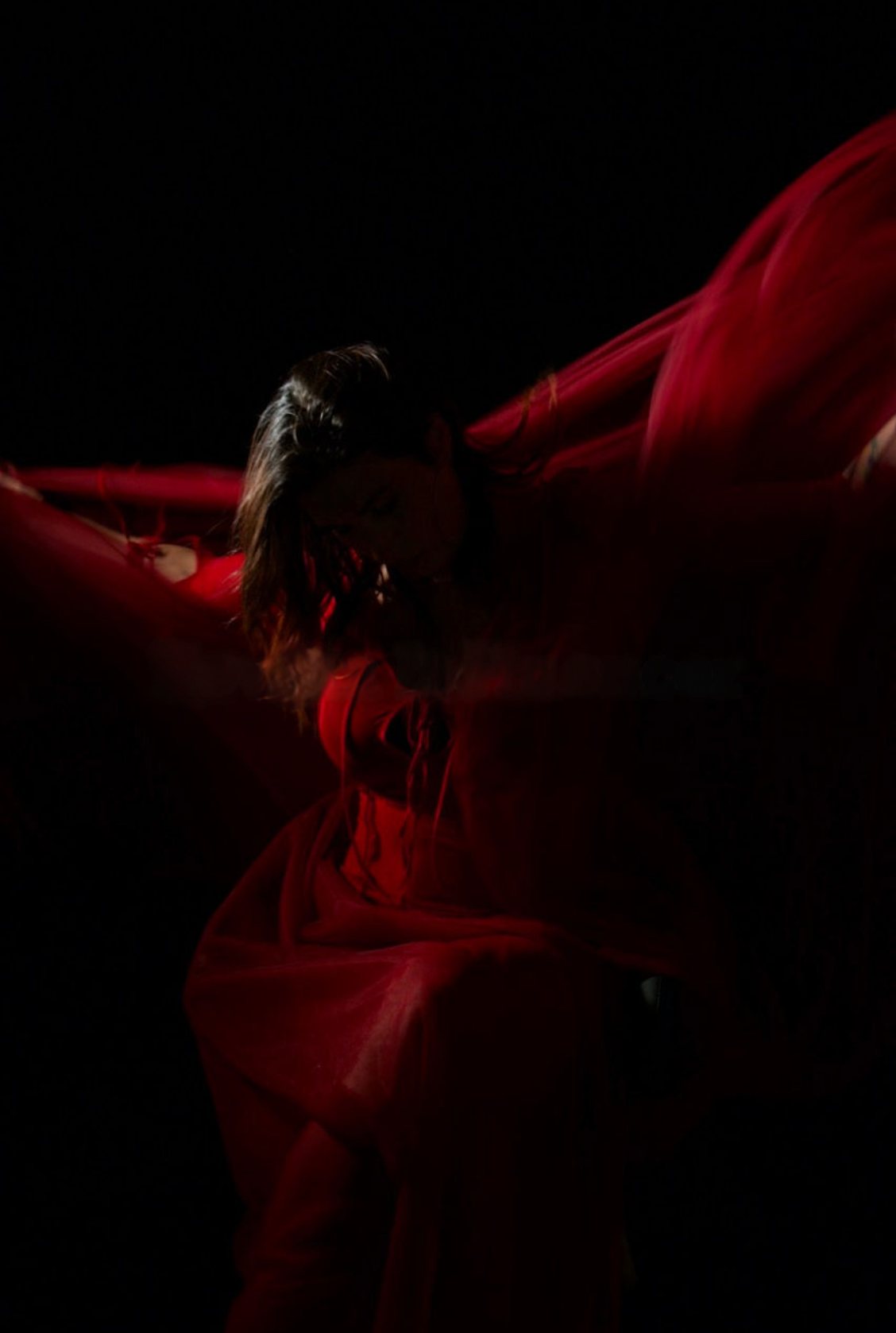 Ophelia envuelta en tela roja en movimiento, fondo oscuro. Vertical.